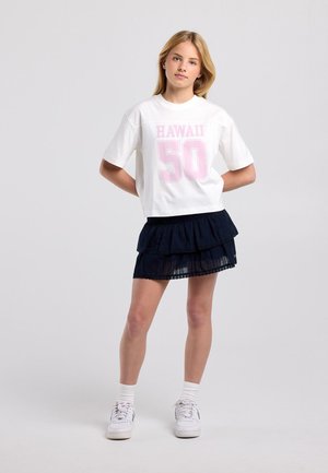 Junges Mädchen steht nach vorne gerichtet, trägt ein weißes "HAWAII 50"-T-Shirt, einen schwarzen Stufenrock, weiße Socken und weiße Sneaker vor schlichtem Hintergrund.