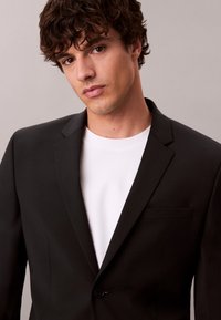 Νέος άνδρας με σγουρά σκούρα μαλλιά, ντυμένος με μαύρο σακάκι πάνω από ένα λευκό μπλουζάκι, κοιτάζει ελαφρώς πλάγια σε απλό φόντο.