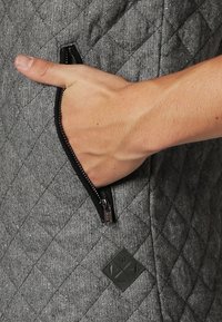 Main insérée dans la poche dézippée d'un vêtement en tissu matelassé gris avec une fermeture éclair noire visible et un petit patch noir.