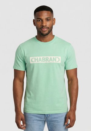 Uomo che indossa una T-shirt verde menta a maniche corte con la scritta "CHABRAND", in piedi davanti a uno sfondo grigio chiaro uniforme.