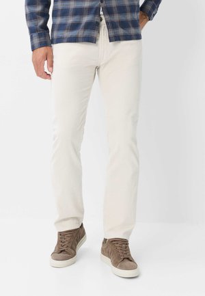Uomo con pantaloni beige, sneaker in pelle scamosciata marrone e camicia a quadri blu e marrone con una mano in tasca.