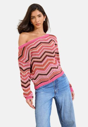 Frau trägt einen schulterfreien Pullover mit Zickzackmuster in Pink, Braun und Orange, langen Ärmeln und hellblauen, hoch taillierten Jeans, posiert leicht sich lehnend.