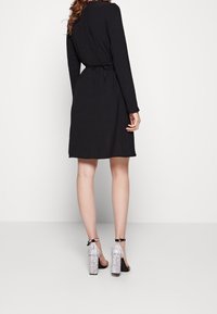 Robe longue noire à manches longues avec une taille cintrée et un détail noué, associée à des chaussures à talons blocs argentées pailletées. Texture de tissu lisse.