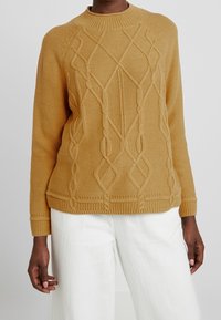 Pull en tricot jaune moutarde avec un motif torsadé texturé, encolure ronde et poignets côtelés. Associé à un pantalon blanc.