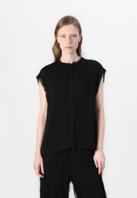 Svart ermeløs topp med frynser langs halsringningen og armhulene. Enkel design med en rett silhuett og myk tekstur.