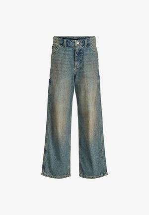 Personne portant une chemise à carreaux noir et blanc par-dessus un t-shirt blanc, un jean ample délavé et des bottes beige, debout sur un fond uni.