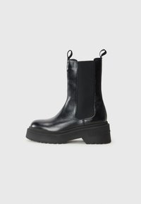 Bottine noire avec panneaux élastiques sur les côtés et languettes de traction, dotée d'une semelle épaisse portant la mention "TOMMY". Design simple et épuré sur fond blanc.