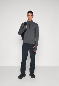 Gray long-sleeve zip-up shirt, black pants with knee seams, and black shoes. Person holding a black backpack, standing against a plain background.