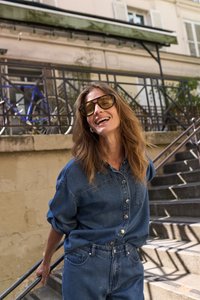 Femme portant de grandes lunettes de soleil et une tenue en jean, souriante, tenant une rampe sur des marches en pierre en extérieur dans un cadre urbain ensoleillé.