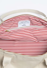Interieur van een canvas tas, met een ritscompartiment met een rood-wit gestreepte voering. Gouden rits en label met verzorgingsinstructies zichtbaar.