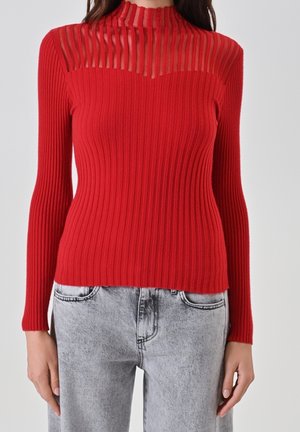 Femme portant un pull rouge moulant à côtes avec un empiècement rayé transparent et un col montant, associé à un jean gris clair délavé à l'acide.