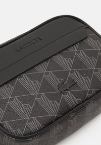 Borsa rettangolare nera Lacoste con nome del marchio in rilievo, motivo geometrico e piccolo logo coccodrillo su superficie testurizzata.