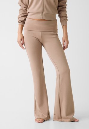 Mujer con pantalones acampanados beige y chaqueta a juego con cremallera, de pie descalza frente a un fondo claro y liso.