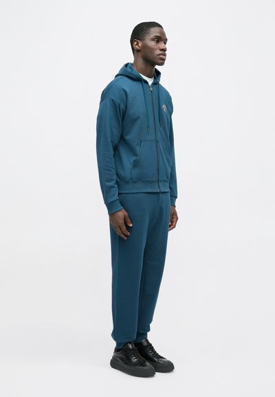 Modrá mikina s kapucňou, predným vreckom, zipsom a šnúrkami, doplnená o zladené zužujúce sa tepláky. Čierne tenisky dopĺňajú outfit.