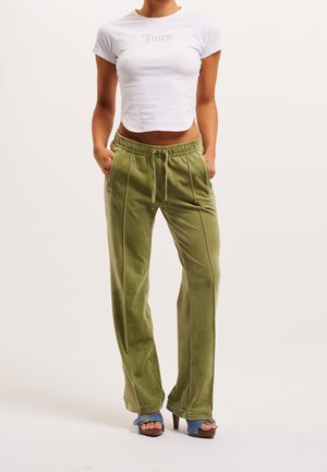 Frau trägt ein eng anliegendes weißes Crop-Top mit dem Text "Juicy", olivgrüne Hosen mit Kordelzug und blaue Sandalen mit Absatz, steht mit den Händen in den Taschen.