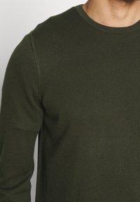 Pull en maille vert olive avec un col rond, présentant une texture côtelée, des manches longues et des détails sur les coutures latérales.