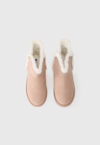 Svetlohnedé slip-on papuče s nadýchanou bielou kožušinou a stehom pozdĺž podrážky, vystavené vedľa seba na bielom pozadí.