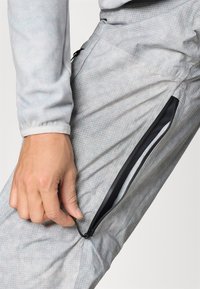 Gaišie pelēkie bikses ar režģa rakstu, sānu rāvējslēdzēja kabatu ar melniem akcentiem un teksturētu apdari, kas nodrošina vieglu kustību.