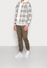 Kostkovaná flanelová košile v bílé, béžové a šedé barvě přes bílou mikinu s kapucí. Outfit doplňují olivové cargo kalhoty a bílé tenisky.