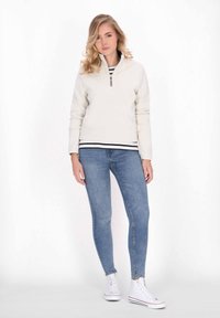 Fleece zip-up color crema chiaro con maniche lunghe, indossato sopra una maglietta a righe bianche e nere, abbinato a jeans skinny blu e sneakers bianche.