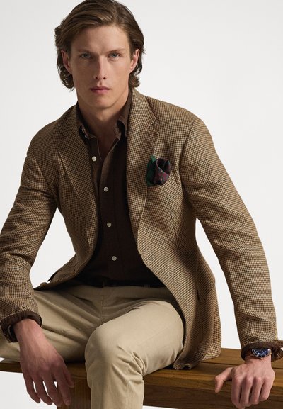 Blazer marrom com padrão pied-de-poule sobre uma camisa escura, combinado com calças chino bege. Detalhes notáveis incluem um lenço de bolso estampado e um relógio.