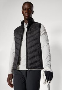 Coletes acolchoados pretos com design acolchoado, camisa polo de manga longa cinza e calças de golfe pretas; uma mão en luva segura um taco de golfe.