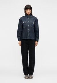 Πουκάμισο από τζιν σε σκούρο μπλε με δύο τσέπες στο στήθος, κλασικό γιακά, κούμπωμα με κουμπιά και αντίθετες ραφές, συνδυασμένο με μαύρο παντελόνι φαρδιάς γραμμής.