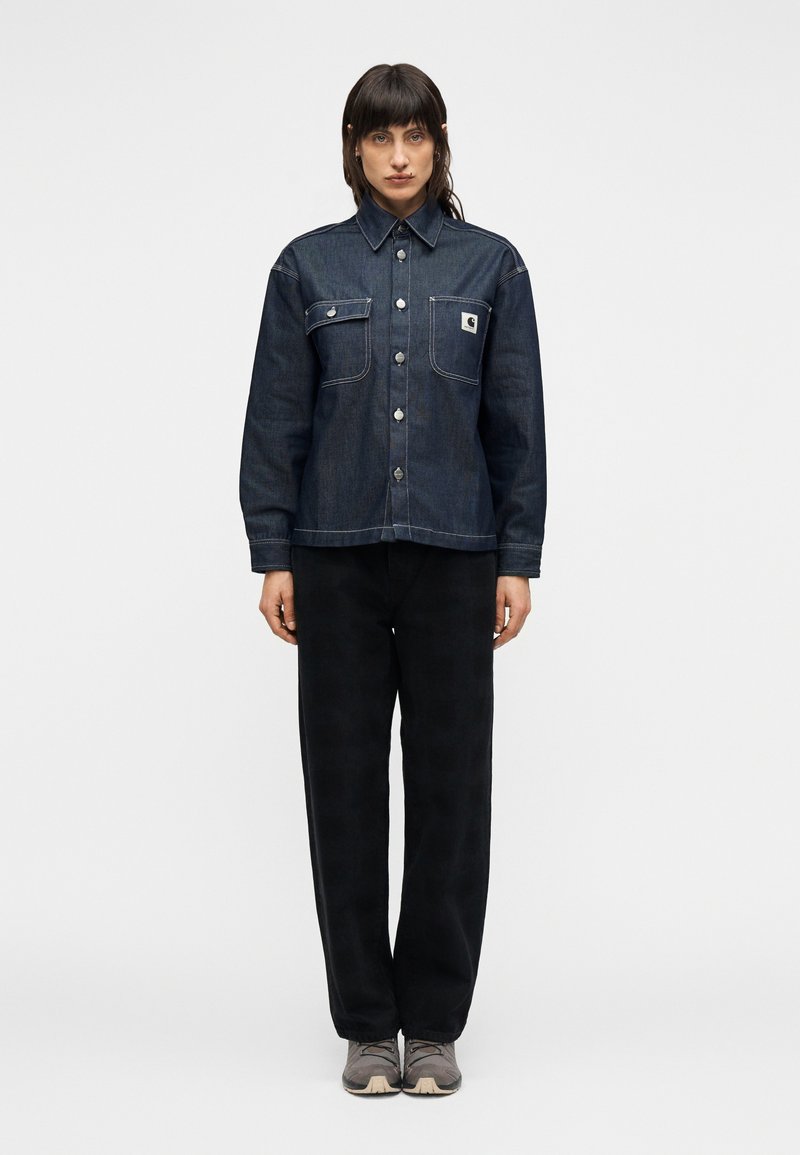 Πουκάμισο από τζιν σε σκούρο μπλε με δύο τσέπες στο στήθος, κλασικό γιακά, κούμπωμα με κουμπιά και αντίθετες ραφές, συνδυασμένο με μαύρο παντελόνι φαρδιάς γραμμής.