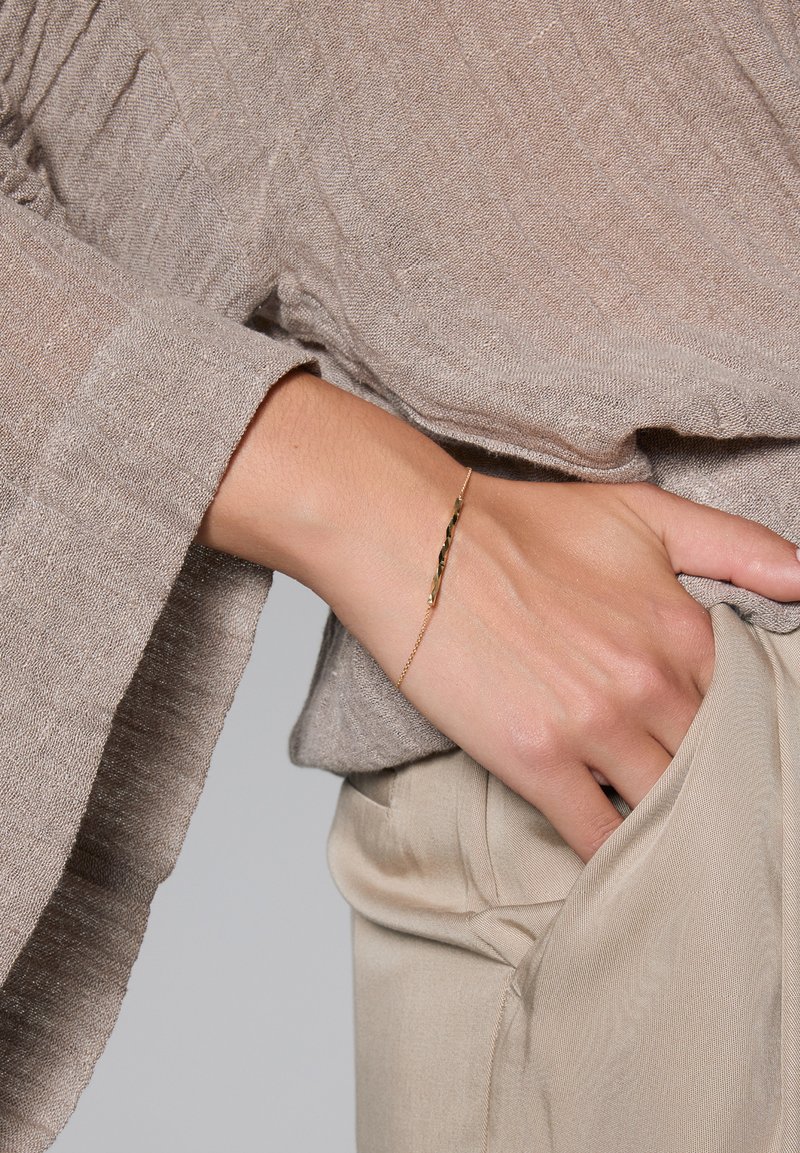 Złoty skręcony bransoletka na nagim nadgarstku, w połączeniu z jasnobeżową, teksturowaną bluzką i dopasowanymi spodniami, prezentując swobodną elegancję.