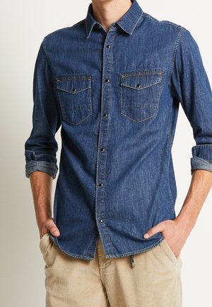 Homme portant une chemise en denim bleue à manches longues avec des poches poitrine et un pantalon beige en velours côtelé, mains dans les poches, manches retroussées.