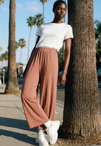 Egy fehér pólót, bő rózsaszín nadrágot és fehér sportcipőt viselő nő dől egy pálmafához egy napsütéses járdán, amelyet pálmafák szegélyeznek.