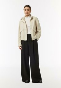 Beżowa krótka kurtka z dwiema przednimi kieszeniami i zamkiem, w połączeniu z czarnymi spodniami typu wide-leg. Model nosi białą bluzkę z golfem pod spodem.