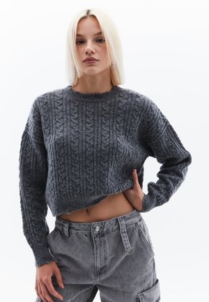 Grauer, verkürzter Pullover mit einem Zopfmuster, Rundhalsausschnitt und langen Ärmeln, kombiniert mit hochgeschnittenen grauen Hosen mit strukturierten Details.