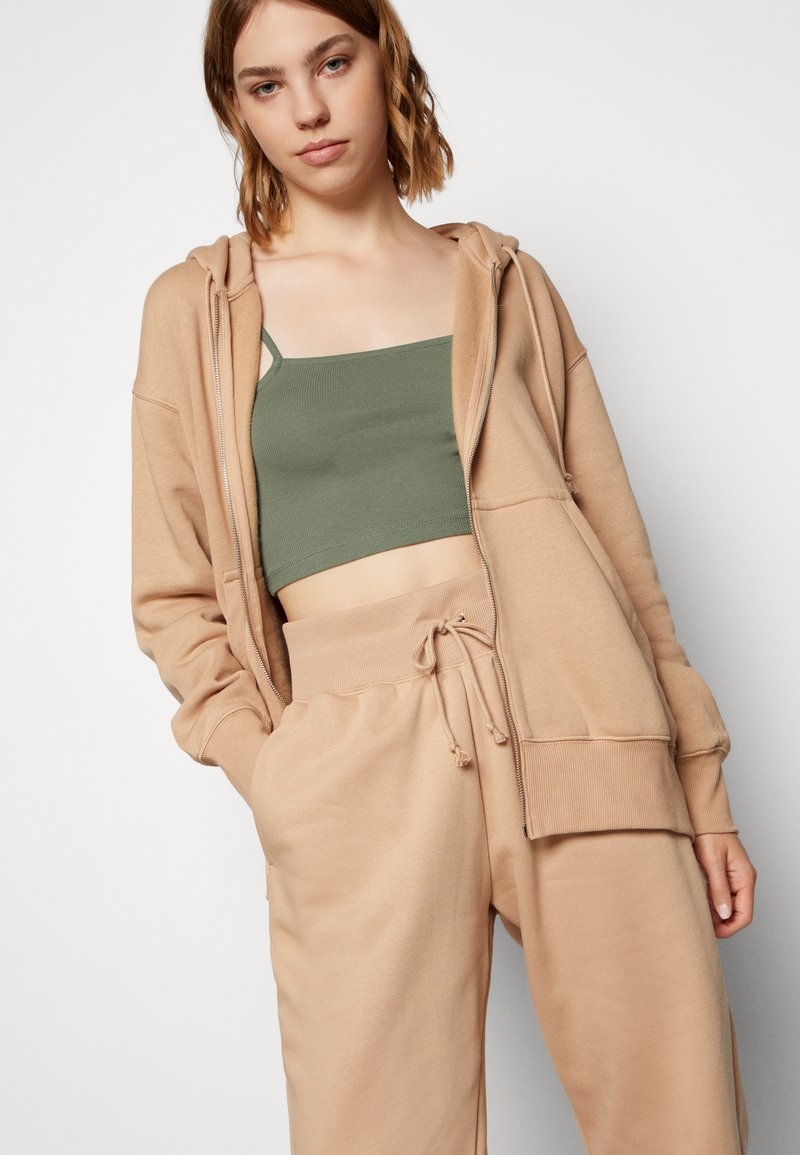 Felpa beige con zip e tasche frontali, abbinata a un top corto verde e pantaloni della tuta beige con cordino. Tessuto morbido e testurizzato.