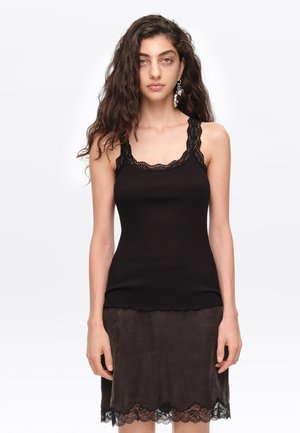 Mujer con cabello largo y rizado que lleva una blusa negra sin mangas con ribete de encaje y una falda oscura con bajo de encaje, de pie sobre un fondo blanco.