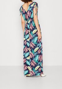 Kobieta w granatowej sukience maxi w kolorowy tropikalny wzór liści, z dekoltem w kształcie litery V, krótkimi rękawami związanymi, oraz białych szpilkach.