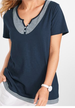 Kvinde iført navyblå kortærmet top med stribet V-udskæring og kant, hvide bukser og hvidt armbåndsur, beskåret ved skuldre og hofter.