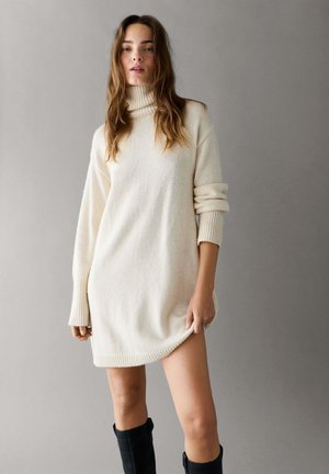 Mujer de pie con un vestido de punto color crema con cuello alto y botas negras hasta la rodilla sobre un fondo gris liso.