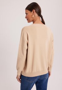 Beżowy sweter dziany o luźnym kroju, z obniżonymi ramionami i ściągaczami. Połączony z niebieskimi dżinsami o gładkiej fakturze i minimalnych detalach.