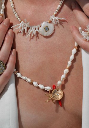 Deux colliers superposés présentent des coquillages, des formes de corail et des accents dorés. Le premier inclut une coquille Saint-Jacques avec une pierre précieuse, tandis que le second a un charme en forme d'œil.