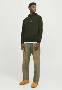 Mørkegrøn hættetrøje med et logo i lys orange, parret med beige brede jeans og brune arbejdssko. Simpelt design, afslappet stil.
