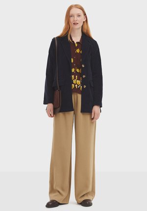 Blazer in velluto a coste blu navy con bottoni dorati, indossato sopra una camicia a fantasia marrone e pantaloni beige a gamba larga, abbinati a scarpe in pelle scura.