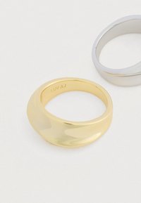 Dva hladké, zakrivené prúžky, jeden zlatý s gravírovaním "LUV AJ" vo vnútri a jeden strieborný, umiestnené na svetlom povrchu.