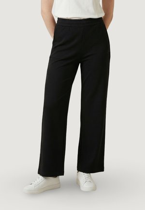 Persona che indossa pantaloni neri a gamba larga con elastico in vita e sneakers bianche, in piedi contro uno sfondo chiaro e uniforme.