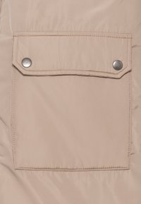 Beige, vadderad ytterklädsel med en stor, rektangulär ficka säkrad med två silverfärgade tryckknappar, synligt sömnad och slät yta.