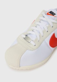 Valged tennisetossud punase Nike logoga; omaduseks tekstuurne samet ja sile kangas, nööriga disain ning metallist silmade aksent.
