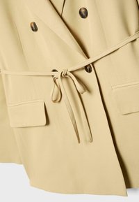 Bež dvojno zapet blazer s temnimi gumbi, dvema sprednjima žepoma s poklopcem in tankim pasom, ki je zavezan v pentljo v predelu pasu.