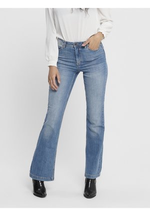 Kobieta w jasnoniebieskich jeansach bootcut, białej bluzce z długim rękawem, z jedną ręką w kieszeni i czarnych botkach.