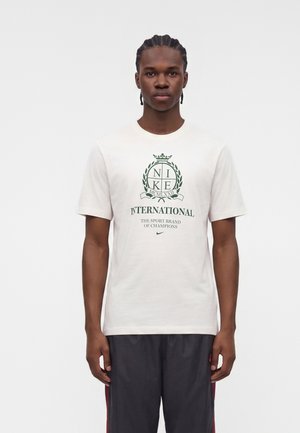 Joven con trenzas que lleva una camiseta blanca de Nike con logo y texto en verde, de pie con los brazos relajados sobre un fondo blanco liso.