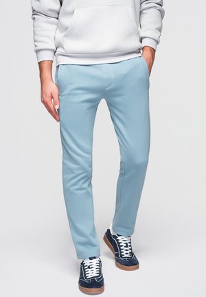 Persona che indossa pantaloni aderenti azzurri, felpa bianca con cappuccio e sneakers blu navy con lacci bianchi, in piedi davanti a uno sfondo uniforme.